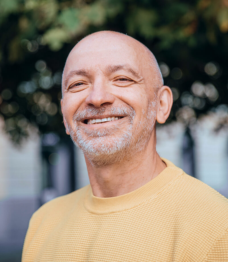 Smiling bald man with a gray beard wearing a yellow sweater outdoors.