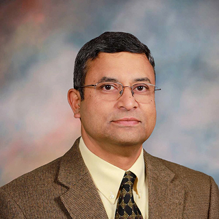 Professional headshot of a male physician wearing glasses, a suit jacket, and tie against a studio background.
